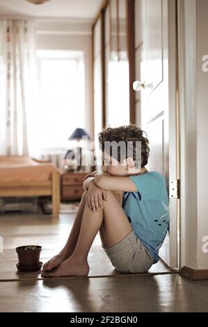 A little boy sits on the floor in a doorway in a room. The child is sad and very upset. Stock Photo