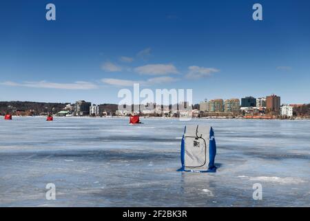 Barrie, Ontario, Canada - March 7, 2021: Ice fishing huts on frozen Kempenfelt Bay of Lake Simcoe in winter with cityscape of Barrie Stock Photo