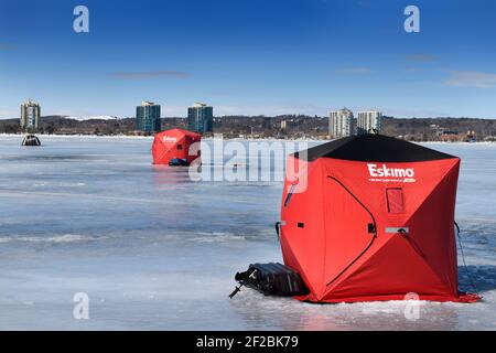 Barrie, Ontario, Canada - March 7, 2021: Red ice fishing tent on frozen Kempenfelt Bay of Lake Simcoe in winter with Barrie condos and landfill hill Stock Photo