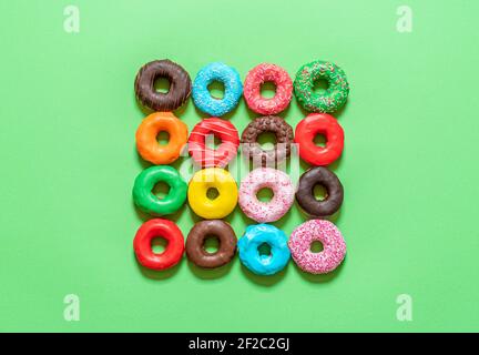 Multicolored icing doughnuts arranged symmetrically in a square on a green background. Homemade chocolate donuts decorated with sprinkles and colored Stock Photo
