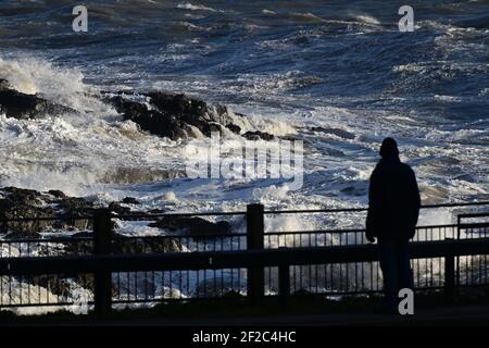Strong Winds and Storm Surf batter the coastline at Mumbles Head. A walker is stopped in his tracks in awe of the power of the waves. Stock Photo