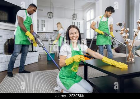 Pretty smiling Caucasian woman in green apron and yellow gloves, wiping table with microfiber cloth and detergents and smiling to camera. Cleaning