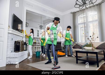 Handsome young african worker of cleaning service, cleaning up carpet and dancing, listening to music in headphones, while another members of cleaning Stock Photo