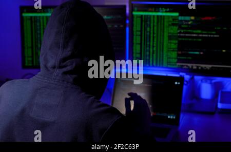 man in a hood working as a hacker at the computer in the dark room at night, hacking the system and laundring secret information. Stock Photo