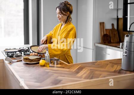 Happy woman dressed in yellow bathrobe cooks pancakes for breakfast, while standing near the hob in the modern kitchen interior at home Stock Photo