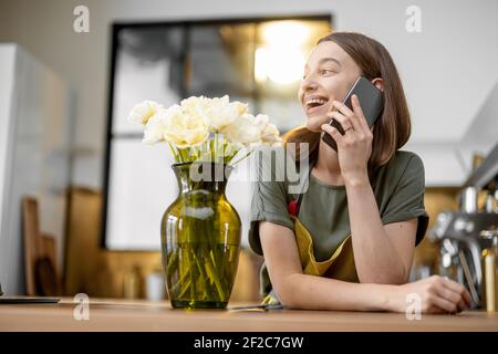 Young woman speaks on phone while staying near a bouquet of fresh tulips in stylish kitchen. Florist with flowers on the kitchen at home Stock Photo
