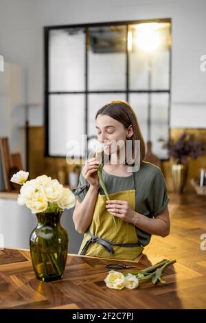 Young woman woman inhales the aroma of fresh tulips in green apron while decorates home interior with a bouquet of fresh flowers. Florist with flowers on the kitchen at home.  Stock Photo