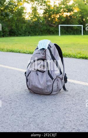 School backpack with medical mask on the ground, green soccer field and sunset background. Back to school during epidemic concept. Stock Photo