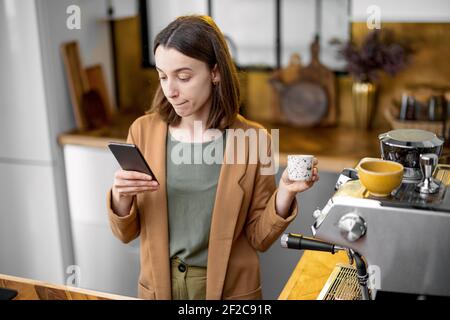 Young stylish woman drinking coffee before going to work in the morning on the beautiful kitchen and chatting on phone. Stock Photo