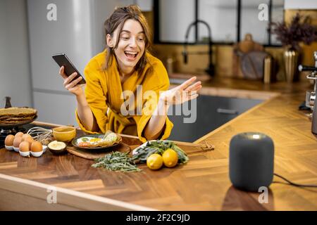 Happy woman in bathrobe controlling home devices with a voice commands, speaking to a smart column during breakfast time on the kitchen at home. Smart home concept Stock Photo
