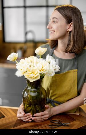 Young woman in green apron with a bouquet of fresh tulips staying on the stylish kitchen. Florist with flowers on the kitchen at home Stock Photo