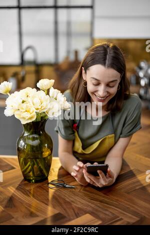 Positive young housewife using smartphone near bouquet of fresh tulips on stylish interior of kitchen. A vase with flowers on wood table. Stock Photo