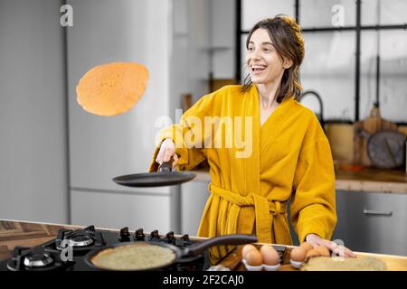 Happy woman cooks pancakes for breakfast, having fun throwing up fried pancakes on the kitchen. Good morning and cooking at home concept Stock Photo