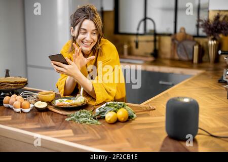 Happy woman in bathrobe controlling home devices with a voice commands, speaking to a smart column during breakfast time on the kitchen at home. Smart home concept Stock Photo