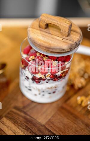 Healthy breakfast with granola made in glass jar with raspberry and organic yogurt on wooden table top in kitchen with nuts on background. Close up Stock Photo