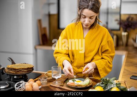 Young woman dressed in yellow bathrobe preparing eggs for breakfast, putting greens on a plate on the kitchen at home Stock Photo