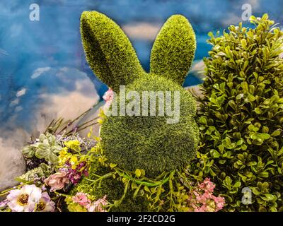 Decorative green topiary rabbit head with flowers around its neck and surrounded by greenery in front of blue sky-like background - Great for spring o Stock Photo