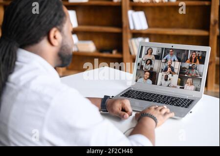 Back view on the laptop screen with many profiles on it, an African-American guy is using computer app for video meeting, online conference with coworkers, colleagues, virtual briefing Stock Photo