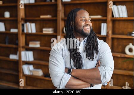 Smiling African office employee with dreadlocks in smart casual wear stands with arms crossed in contemporary coworking office space, a multiracial guy looks away and laughs. Diverse work team concept Stock Photo