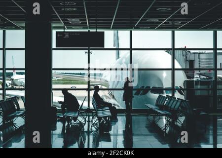 Three persons are wainting in an airport lounge for their departure. A big plane is stainding outside. Stock Photo