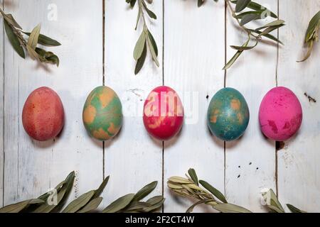 Two Easter symbols: eggs and olive twigs. Five coloured eggs with olive twigs around them on a shabby chic wooden table. Stock Photo