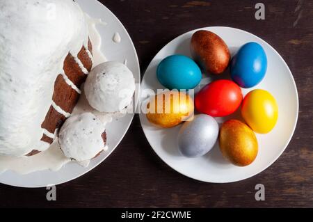 Homemade craft Easter cakes covered with white icing and multicolored painted Easter eggs on plates on a dark wooden background. Stock Photo