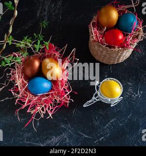 Homemade multicolored painted Easter eggs on multicolored straw in wicker baskets and in stands on a dark background, Easter decoration, top view Stock Photo