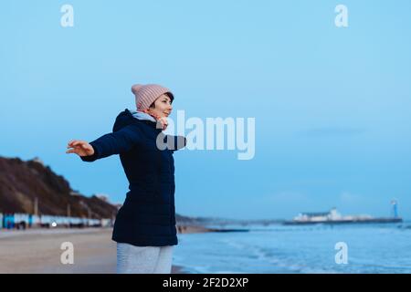 Woman in warm clothes practicing balance and enjoying the moment and sea view during a walk on the coast. Relax and personal fulfillment. Travel in au Stock Photo