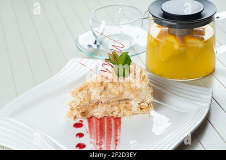 Napoleon cake decorated with a spiral of red chocolate, mint and berry jam on a white plate against a background of orange tea in a teapot and a trans Stock Photo