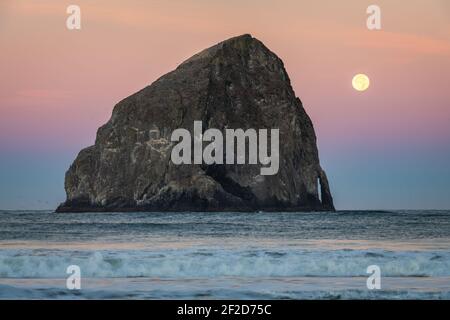 The full moon sets in the dawn light behind Haystack Rack in Oregon Stock Photo