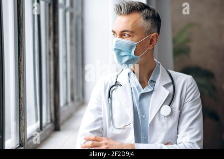 Male medical spezialist, middle-aged caucasian doctor, in a medical uniform and a protective mask, stands in a clinic, looks pensively out the window Stock Photo