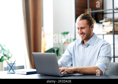 Confident successful guy, freelancer or employee, sits at his work desk at home or in the office, uses a laptop, chatting with colleagues, responding to emails, browsing the Internet Stock Photo