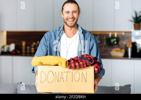 A friendly smiling candid caucasian male volunteer holds a cardboard box with a donation, for reuse or recycle of old clothes. Satisfied handsome man holding a donation box for people in need Stock Photo
