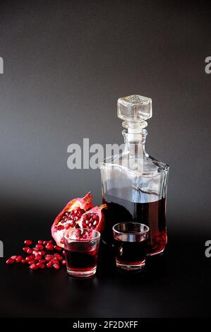 Pomegranate liqueur in a bottle and two glasses, next to the seeds and fruit on a black background. Vertical positioning. Stock Photo