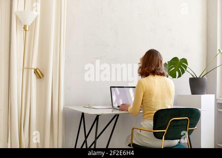 Back view of unrecognizable female in smart casual clothes sitting at desk and browsing data on netbook with empty screen while working in stylish wor Stock Photo