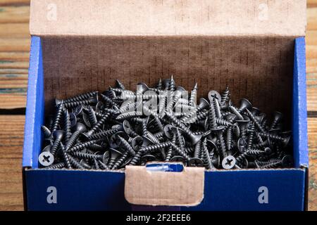 Black screws in a cardboard box. Wooden texture background. Screws for plasterboard. Stock Photo