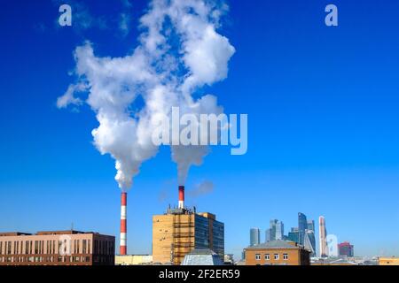 White puffs of smoke rise into the blue cloudless sky from industrial chimneys above the rooftops of houses in the city. Sunny frosty day in winter. Stock Photo