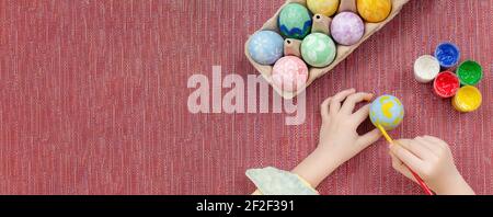 A boy painting an Easter egg, top view on a red fabric background. Painting Easter eggs, gouache paints, brush, preparation for the holiday Easter. Ba Stock Photo
