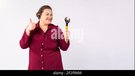 Banner, long format, grey background Female fat mechanic or a plumber, showing thumb up, working hard, holding a wrench, wearing bordo dress and Stock Photo
