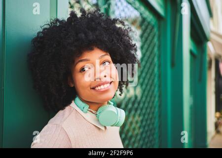 Smiling afro-haired woman with headphones leaning against green wall. Portrait of happy latina woman . High quality photo Stock Photo