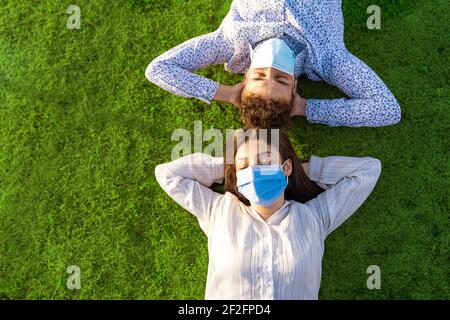 Happy couple despite social distancing due to Coronavirus pandemic. Top view of two young real people resting in the grass of city park wearing protec Stock Photo
