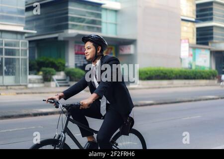 Asian businessman in a suit is riding a bicycle on the city streets for his morning commute to work. Eco Transportation Concept. Stock Photo