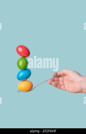 Hand holding a spoon with four easter eggs with different colours. Minimal funny concept with blue background. Stock Photo
