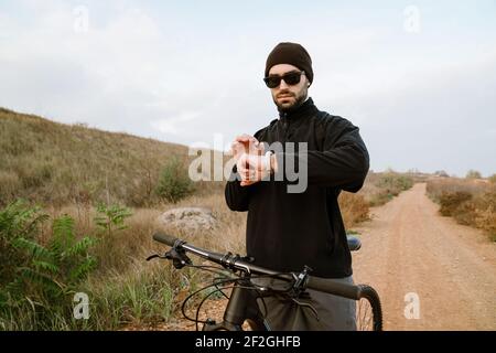 Handsome unshaven guy using smartwatch while having bike ride outdoors Stock Photo