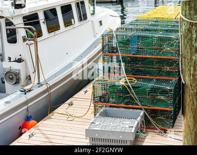 A fishing boat docked next to a wood pier with lobster and crab traps stacked on each other ready to be used. Stock Photo