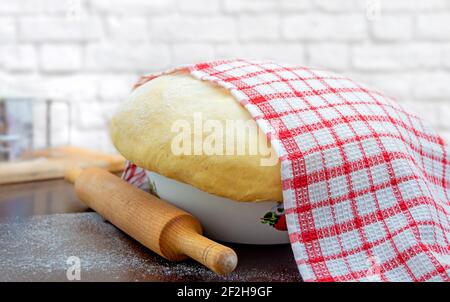 Raw yeast dough covered with a towel in a bowl on the floured kitchen table, recipe idea. Concept home baking or making dough. Stock Photo