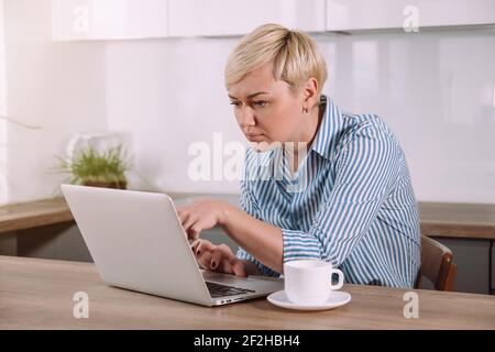 Focused serious woman working on laptop at home Female using portable computer in kitchen Stock Photo
