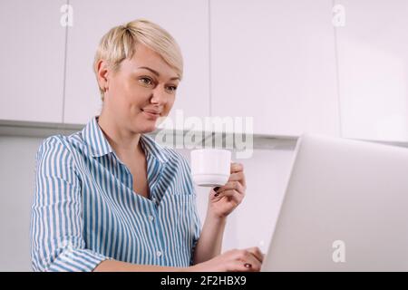 Portrait of a business woman with cup of coffee sitting in kitchen at home near laptop Stock Photo