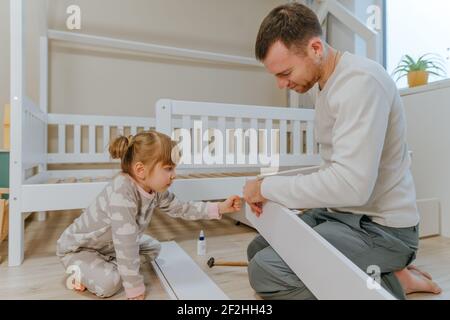 Little 4-years girl helps her father assemble or fixing the drawer of bed in the kids bedroom. Stock Photo
