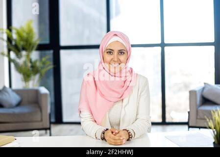 Young educated Muslim Arab female job candidate applicant in hijab sitting at the desk and looking at the camera, feeling grateful and happy to accept the position, holding hands together, and smiling Stock Photo